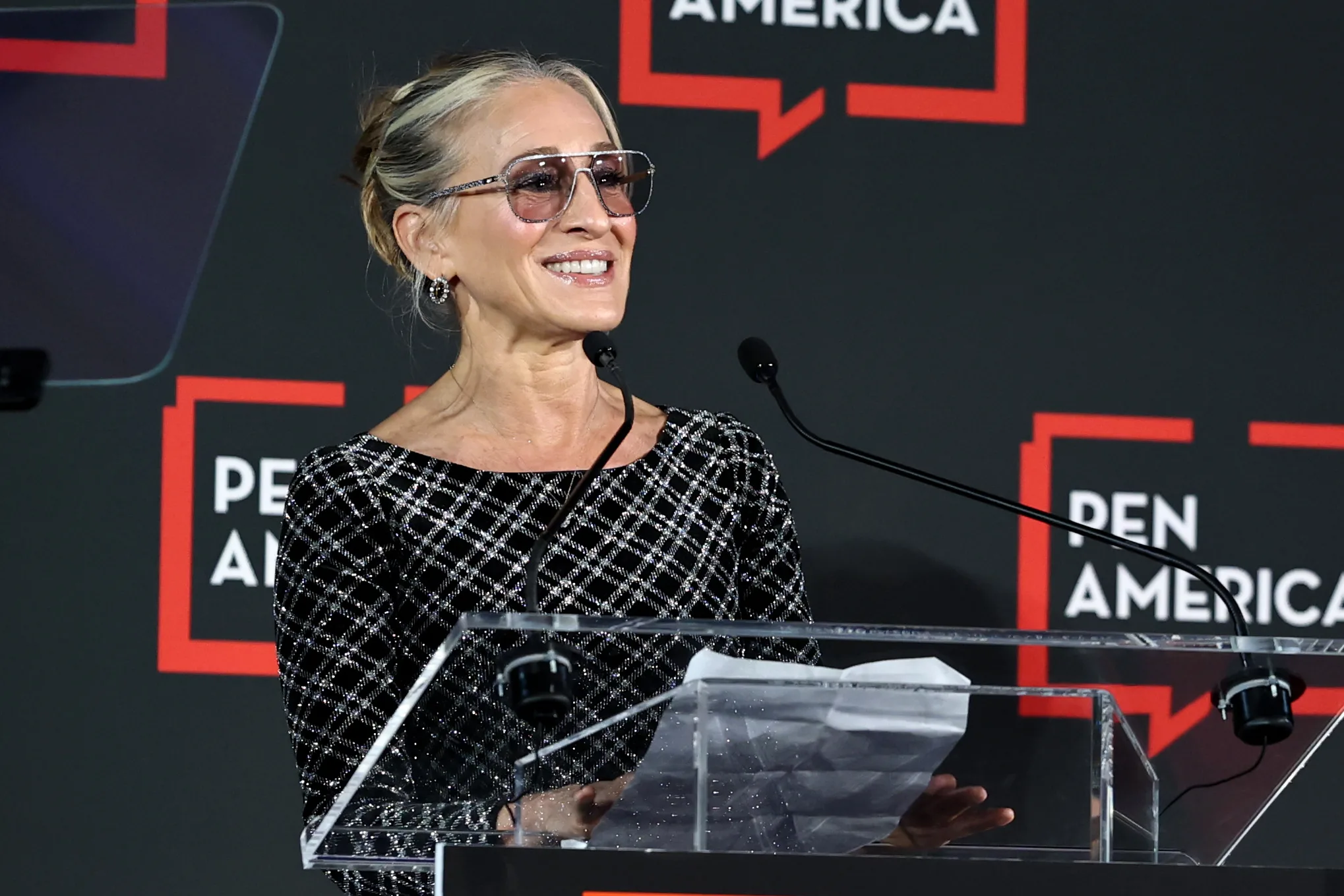 A woman wearing sunglasses and a patterned black dress smiles while speaking at a clear podium, with PEN America logos displayed on a black background behind her.